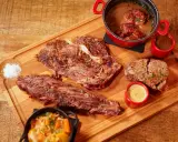 Assortiment de viande à griller, steak, et ragoût de bœuf, servi avec des sauces et des légumes sur une planche en bois.