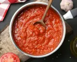 Une casserole remplie de coulis de tomates maison, avec une cuillère en bois. Des ingrédients frais comme du romarin, de l'ail et une tomate coupée en deux sont disposés autour.