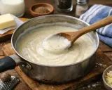 Une cuillère en bois remue une béchamel crémeuse dans une casserole. Ingrédients pour la recette bechamel : lait, beurre, noix de muscade râpée.