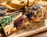 Un gigot d'agneau tendre, rôti à la perfection, est présenté sur une planche en bois avec du romarin frais et des grains de poivre.