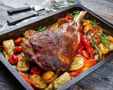 Une succulente épaule d'agneau rôtie dans un plat avec des légumes colorés, des poivrons, des tomates et des herbes aromatiques.