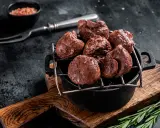 Un plat de rognons de veau grillés, servis dans une cocotte en fonte sur une planche en bois, avec une pincée de sel rose et une branche de romarin.