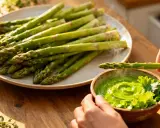 Une main trempe une asperge dans une sauce verte onctueuse, parfaite pour une recette sauce asperges. Des asperges cuites à la vapeur sont sur une assiette à côté.