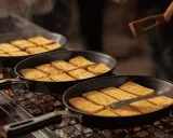 Des tranches de fromage fondent dans des poêles sur un raclette barbecue. Un ustensile en bois retourne délicatement les morceaux dorés.