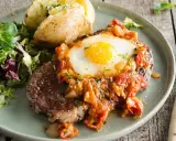 Un délicieux steak a cheval, garni d'un œuf au plat et d'une sauce tomate, servi avec une pomme de terre au four et une salade verte.