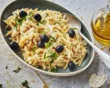 Un plat de bacalhau à brás, effiloché et garni d'olives noires et de persil frais. Une touche d'huile d'olive et d'ail est visible à côté.