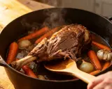 Un tendre agneau de 7 heures mijote dans une cocotte avec carottes, oignons et ail. La viande se détache facilement de l'os, signe d'une cuisson parfaite.
