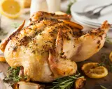 Un délicieux poulet rôti, doré et parfumé aux herbes, est servi sur une planche en bois avec des quartiers de citron et de l'ail rôti.