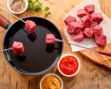 Préparation d'une fondue chinoise : des cubes de viande rouge sur des brochettes trempent dans un bain d'huile chaude. Quel accompagnement fondue viande ? Sauces et épices sont à portée de main.