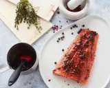 Un filet de saumon mariné est prêt à être cuit sur une planche de cèdre. Des herbes fraîches et des grains de poivre complètent la scène.