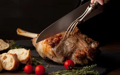 Une main utilise un couteau et une fourchette pour découper un gigot d'agneau rôti, prêt à être servi avec du pain et des tomates.