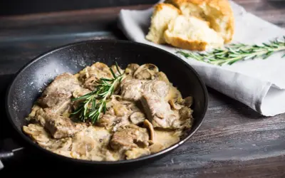 Délicieux sauté de porc crémeux aux champignons, garni de romarin frais, servi dans une poêle avec du pain croustillant à côté.