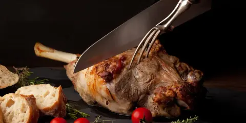 Une main utilise un couteau et une fourchette pour découper un gigot d'agneau rôti, prêt à être servi avec du pain et des tomates.