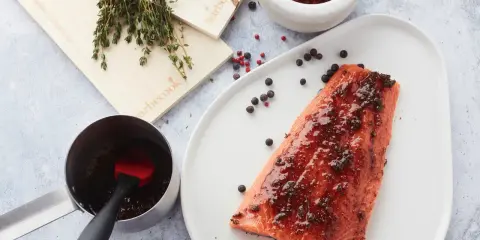 Un filet de saumon mariné est prêt à être cuit sur une planche de cèdre. Des herbes fraîches et des grains de poivre complètent la scène.