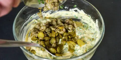 Ajout de câpres et cornichons hachés à une base crémeuse pour préparer une sauce gribiche savoureuse.