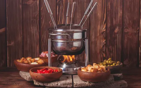 Un appareil à fondue bourguignonne chauffe, entouré de bols de croûtons, d'olives, de tomates séchées et de dés de viande. Idéal pour savoir que faire avec de la viande à fondue bourguignonne.