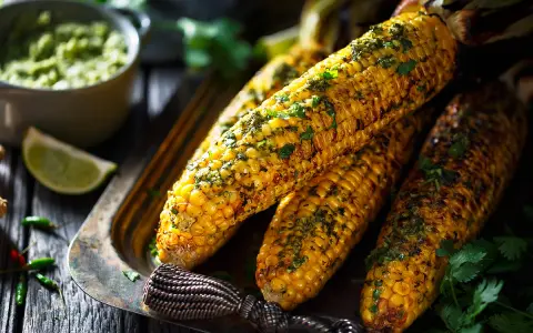 Épis de maïs grillés, parsemés d'herbes fraîches, servis sur un plateau avec une sauce verte et des quartiers de citron vert. Un délice parfait pour un repas mais au barbecue.