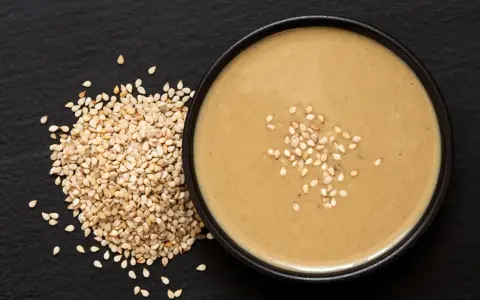 Un bol de sauce tahini onctueuse, garni de graines de sésame, à côté d'un tas de graines de sésame sur fond noir.