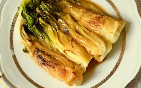 Pak choi braisé, tendre et savoureux, nappé d'une sauce dorée, servi sur une assiette élégante.