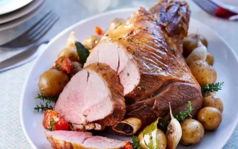 Un gigot d'agneau rôti, tranché, servi avec des pommes de terre nouvelles, tomates et ail. Parfait pour savoir que faire avec des morceaux de gigot d'agneau.