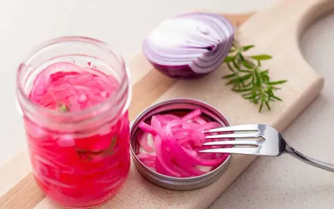 Pickles oignon rouge vibrants dans un bocal, avec une moitié d'oignon rouge et une branche de romarin sur une planche à découper.