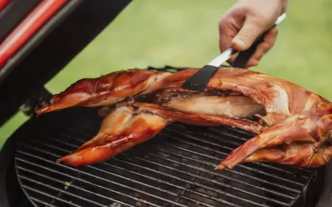 Un lapin entier est cuit sur un barbecue, sa peau dorée et croustillante. Une spatule retourne délicatement la viande pour une cuisson parfaite.