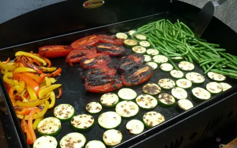 Légumes variés (poivrons, courgettes, tomates, haricots verts) en cours de cuisson sur une plancha. Le temps de cuisson des légumes à la plancha est idéal pour un repas sain et savoureux.