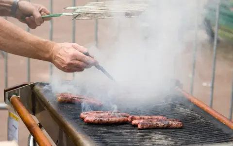 Des merguez grillent sur un barbecue, dégageant une fumée épaisse. Un homme les retourne avec une pince, se demandant s'il faut piquer les merguez pour une cuisson parfaite.