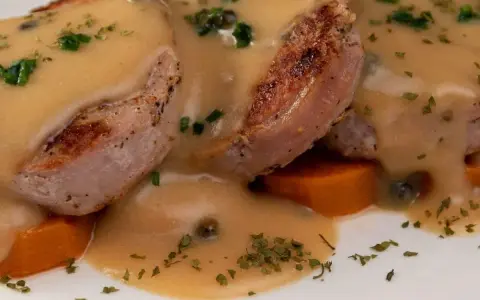 Tranches de gigot d'agneau nappées d'une sauce onctueuse au fond de veau, parsemées de persil et de câpres, sur un lit de carottes.