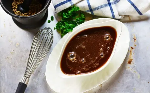 Sauce à la viande fumante dans un saucier, avec un fouet, du persil et un mortier rempli de grains de poivre.