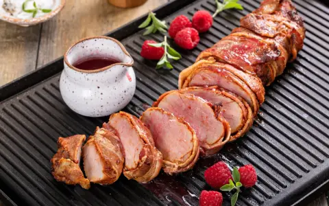 Filet mignon enrobé de bacon, tranché, servi avec des framboises et une sauce rouge sur une plaque de cuisson.