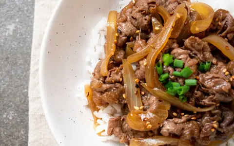 Un plat de riz blanc garni de fines tranches de bœuf aux oignons caramélisés, parsemé de graines de sésame et de ciboule ciselée. Un délice de boeuf aux oignons.