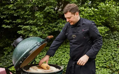Un chef prépare un rôti sur une pierre à pizza dans un barbecue binchotan vert, entouré de verdure luxuriante.
