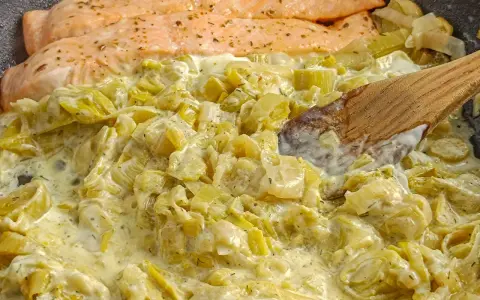 Filets de saumon poêlés sur une fondue de poireaux crémeuse, parsemée d'herbes. Une cuillère en bois remue ce plat savoureux.