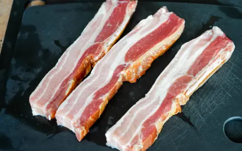 Trois tranches de lard fumé, prêtes pour une recette savoureuse, reposent sur une planche à découper noire.