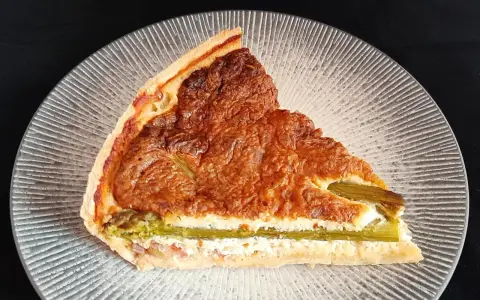 Tranche de tarte aux asperges blanches, garnie d'une préparation crémeuse et dorée, sur une assiette texturée.