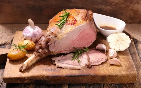 Un gigot d'agneau rôti, tendre et juteux, servi avec des pommes de terre, de l'ail et du romarin sur une planche en bois.