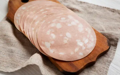 Tranches de mortadelle cuit ou cru, disposées en éventail sur une planche à découper en bois, sur un tissu de lin.