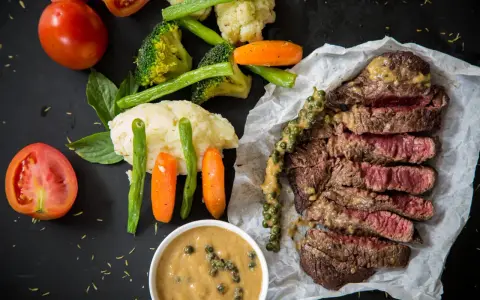 Un steak tranché, servi avec des légumes et deux types de sauces. L'une est une sauce crémeuse aux grains de poivre vert, l'autre est une sauce plus claire dans un bol.
