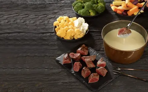 Un morceau de viande est trempé dans un caquelon de fondue. Des morceaux de viande, du brocoli, du chou-fleur et des légumes racines sont disposés sur une table en bois sombre.