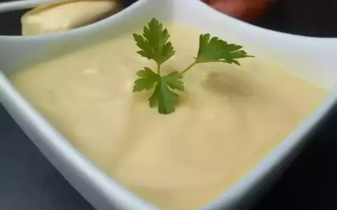 Un bol de beurre blanc crémeux, garni d'une feuille de persil. Des échalotes et un morceau de pain sont visibles en arrière-plan.