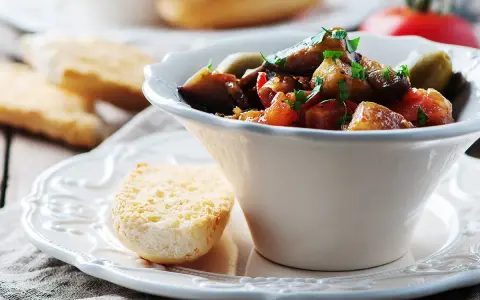 Un bol de caponata sicilienne, plat sicilien aux aubergines, tomates et olives, garni de persil frais. Servi avec du pain grillé.