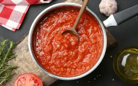 Une casserole remplie de coulis de tomates maison, avec une cuillère en bois. Des ingrédients frais comme du romarin, de l'ail et une tomate coupée en deux sont disposés autour.