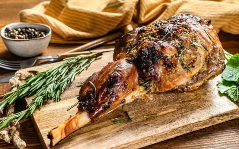 Un gigot d'agneau tendre, rôti à la perfection, est présenté sur une planche en bois avec du romarin frais et des grains de poivre.