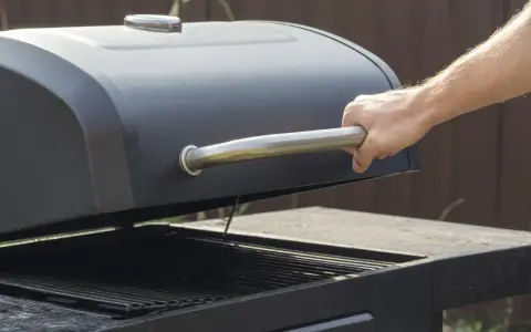 Main d'homme ouvrant le couvercle d'un barbecue noir. Apprenez comment utiliser un barbecue avec couvercle pour des grillades parfaites.