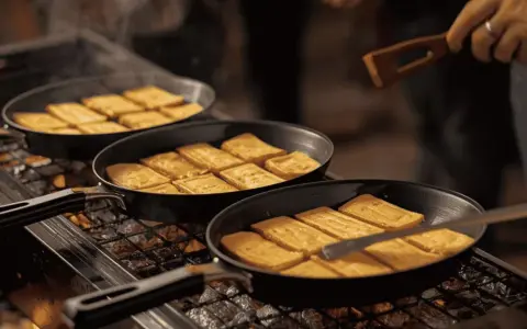 Des tranches de fromage fondent dans des poêles sur un raclette barbecue. Un ustensile en bois retourne délicatement les morceaux dorés.