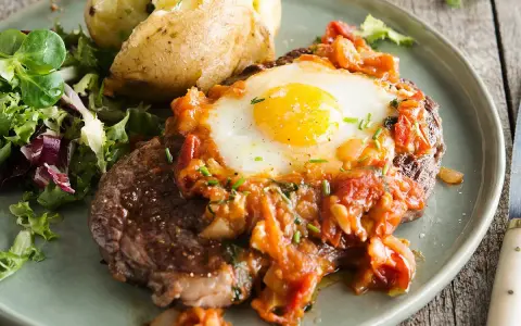 Un délicieux steak a cheval, garni d'un œuf au plat et d'une sauce tomate, servi avec une pomme de terre au four et une salade verte.