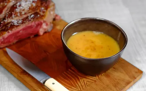 Un bol de sauce pour côte de bœuf, onctueuse et dorée, à côté d'une côte de bœuf saignante et d'un couteau sur une planche en bois.