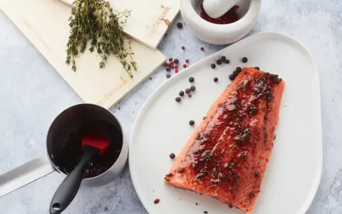 Un filet de saumon mariné est prêt à être cuit sur une planche de cèdre. Des herbes fraîches et des grains de poivre complètent la scène.