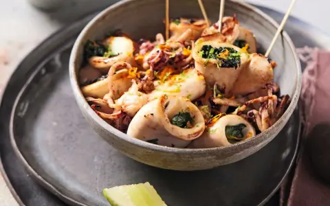 Calamars à la plancha farcis aux herbes, servis dans un bol avec des zestes d'orange et des quartiers de citron vert.